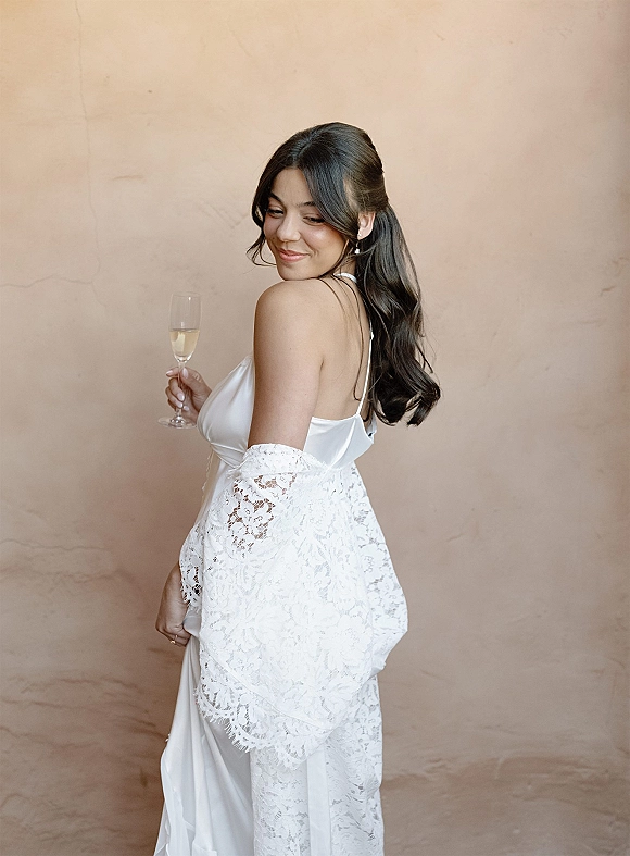 Bridal portrait of a bride holding a champagne glass in a satin slip dress and lace robe, half-up waves against a neutral beige wall