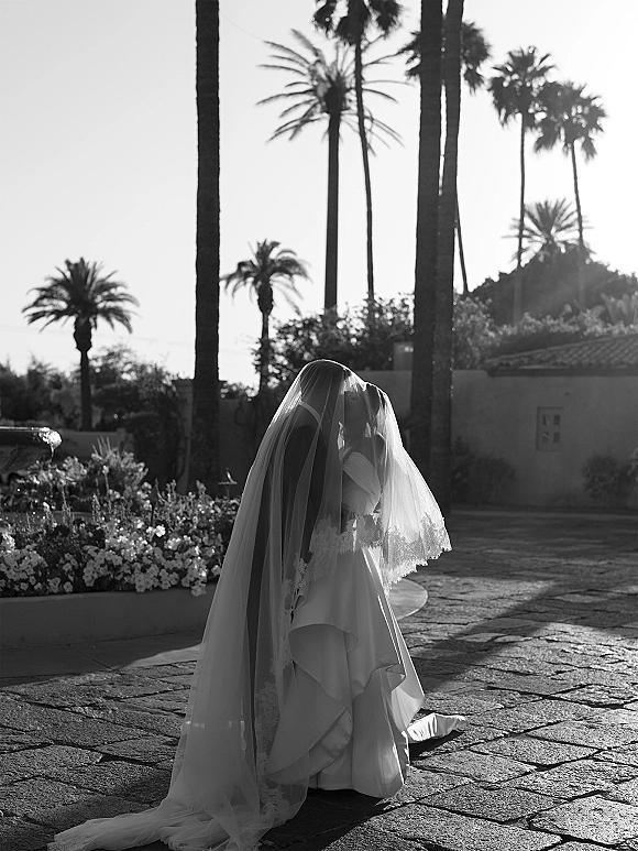 Bridal portrait of a bride in veil walking away, cathedral train trailing on stone courtyard with palm trees, fountain, and flowers behind