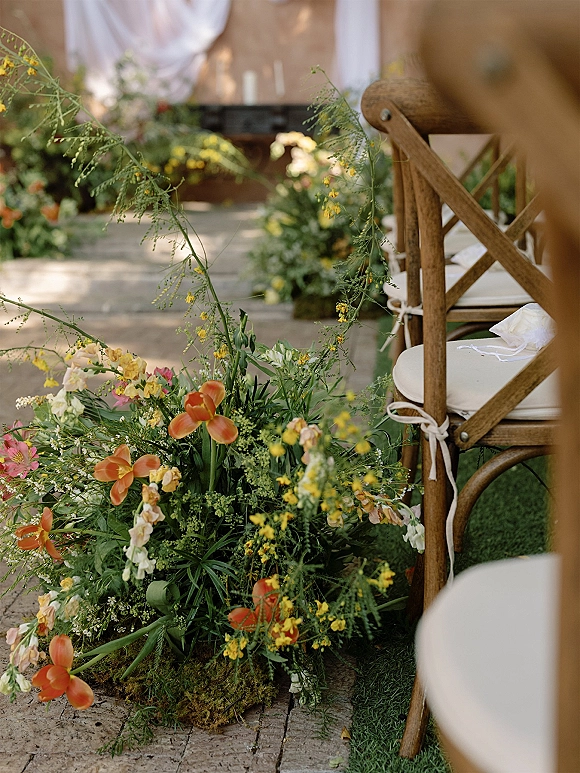 Ceremony aisle decor with aisle floral meadow arrangements of orange and yellow blooms and greenery along a stone aisle with cross back chairs