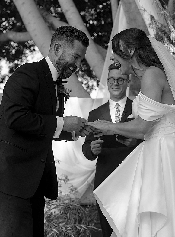 Ring exchange as bride and groom laugh while placing wedding rings, her veil and off-shoulder gown framed by a draped arch and trees