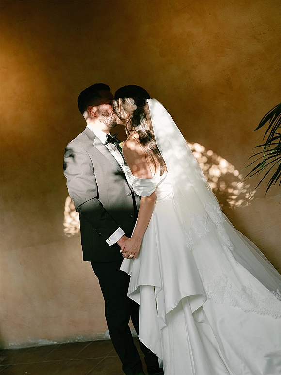 Wedding kiss portrait of bride and groom kissing, holding hands by a stucco wall in dappled sunlight, veil and tuxedo details visible