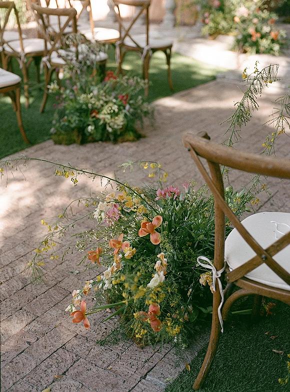 Ceremony aisle decor with aisle ground florals lining a brick pathway, crossback chairs with ribbon ties, and greenery in dappled sunlight