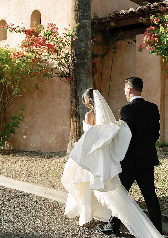 Newlywed couple walking away, bride and groom holding a lace train as a long veil trails by a sunlit stucco villa wall with arches