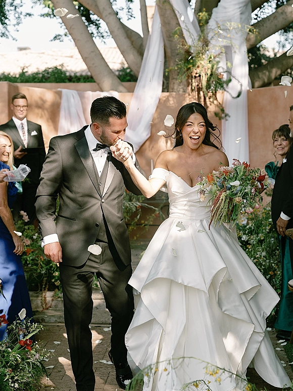 Recessional moment as bride and groom walk the garden aisle, groom kissing her hand while guests toss flower petals under a large tree