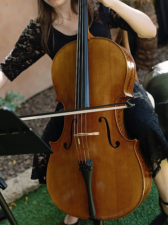Wedding ceremony musician, a wedding cellist in a black lace dress plays cello with bow beside a music stand on a grassy lawn by a stone wall