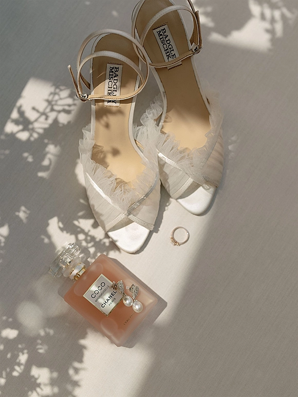 Bridal shoes wedding detail flat lay of white ankle strap heels with tulle trim, perfume bottle, pearl earrings, and ring in window light shadows