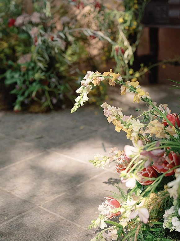 Ceremony aisle flowers in grounded wedding aisle florals with roses, snapdragons, and greenery along a stone floor by a doorway