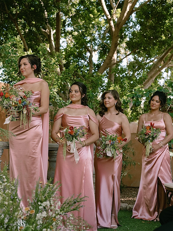 Bridesmaid portraits of bridesmaids in pink dresses, holding bouquets with ribbon wraps by a stone ledge in a green garden setting