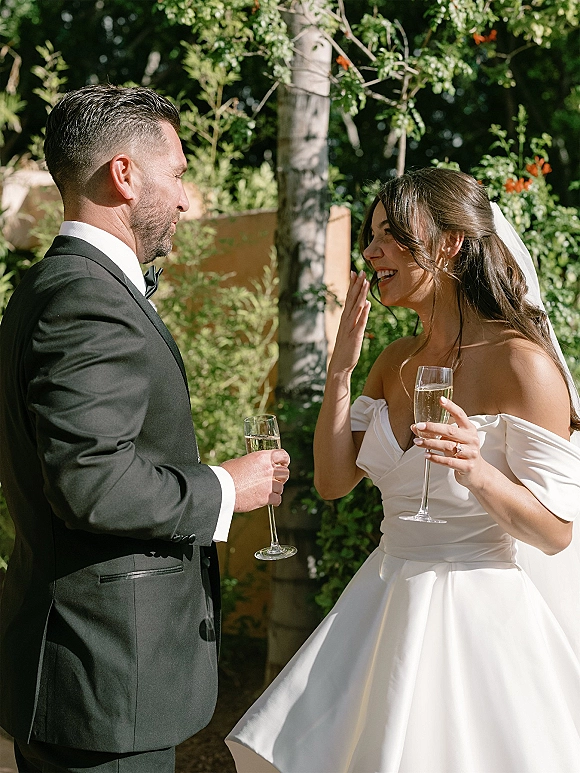 Wedding couple portrait of bride and groom toasting with champagne flutes, her veil and off-shoulder dress glowing in a sunlit garden setting