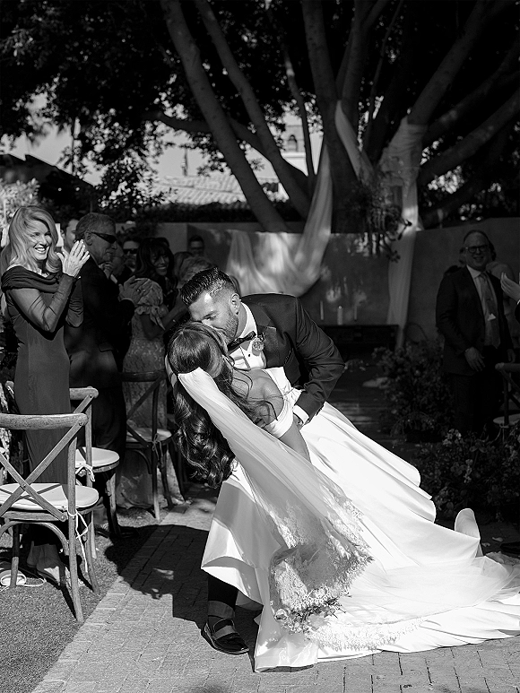 Wedding kiss moment as groom dips bride with long lace-trimmed veil on brick walkway aisle, guests cheering under large trees