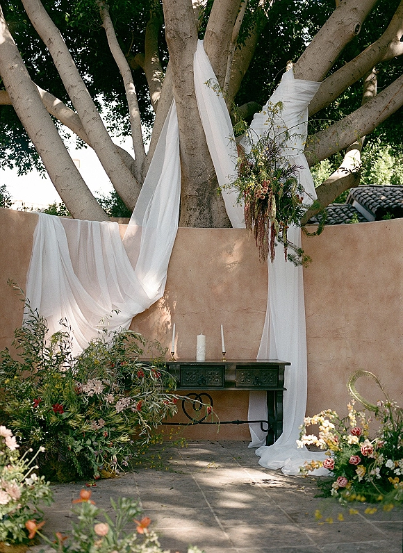 Wedding ceremony backdrop with draped fabric, hanging florals and greenery, plus candles on a console table beneath a large tree in a courtyard