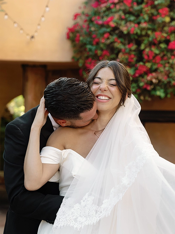 Wedding couple portrait with groom kissing bride’s cheek as she laughs, lace-trim veil on shoulder under string lights by a vine-covered stucco archway
