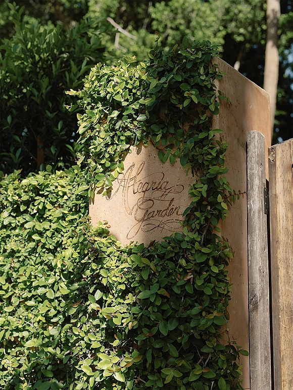 Wedding welcome sign with calligraphy wedding sign lettering on tan stone, framed by greenery vines at a sunlit garden gate entrance