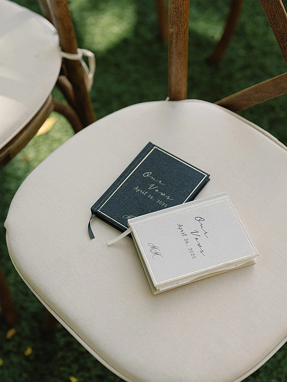 Wedding vow books, his and hers vow books with gold foil lettering and ribbon bookmarks resting on a guest chair on a grassy lawn