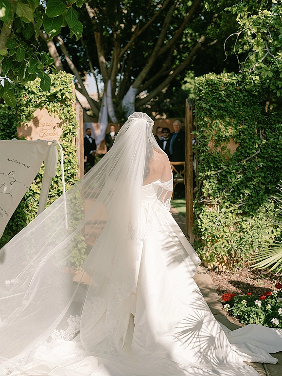 Bridal processional as bride walking down aisle with cathedral veil train and lace trim, entering sunlit garden gate with guests ahead