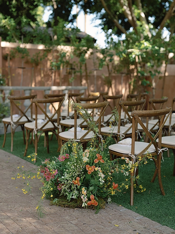 Ceremony aisle decor with cross back chairs and ribbon ties, leading to a colorful ground floral arrangement on a garden walkway