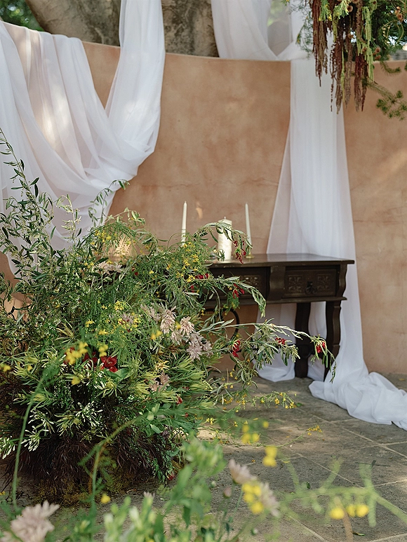 Ceremony backdrop with draped fabric backdrop of white fabric and greenery garland, wildflower florals and taper candles against tan plaster wall