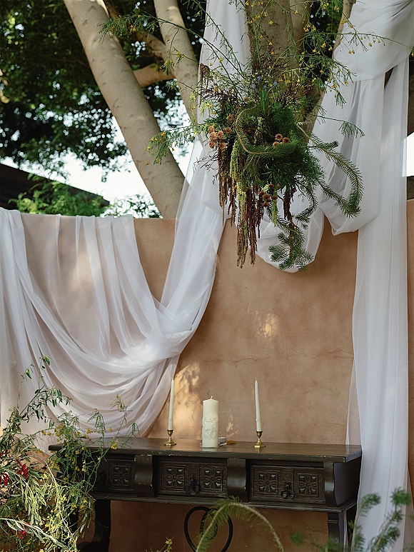 Ceremony backdrop with draped fabric and white chiffon wedding draping, hanging greenery, and brass candles on a dark wood table outdoors