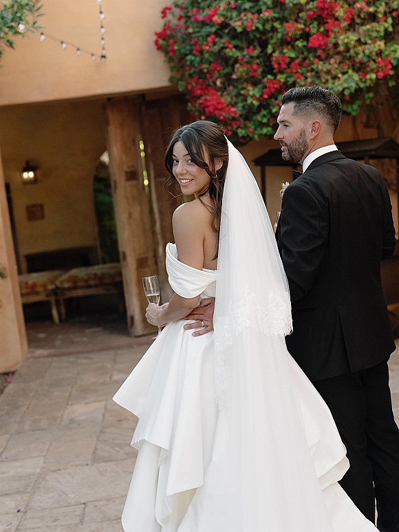 Couple portrait of newlyweds holding champagne as the bride looks back in an off-the-shoulder dress on a string-lit stucco patio