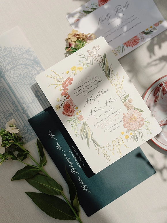 Wedding invitation suite with floral wedding invitations, green envelope, greenery sprigs and small flowers on a white tablecloth flat lay