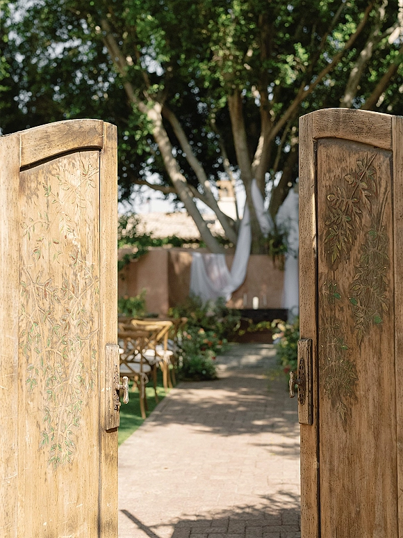 Ceremony entrance with open wooden doors, carved panels and greenery, leading to a candle-lined aisle runner in a courtyard setting