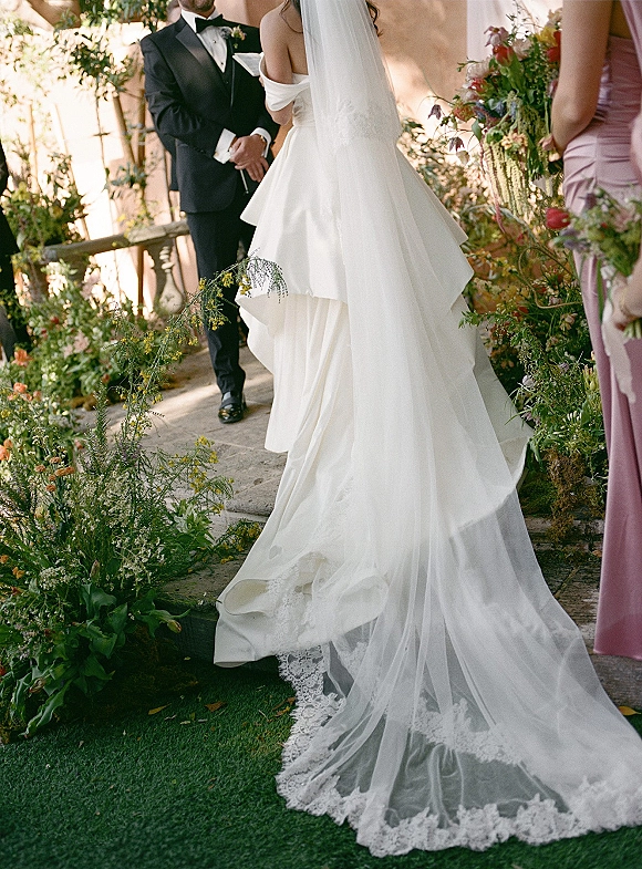 Ceremony moment as bride walking down aisle in cathedral veil and lace train toward groom in tux on stone steps in garden setting