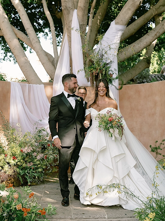 Recessional moment as bride and groom walking hand in hand, she laughs in veil and gown holding bouquet along a stone courtyard aisle