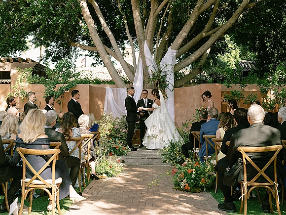 Wedding vows as bride and groom hold hands under a draped greenery arch, brick aisle lined with flowers in a garden setting