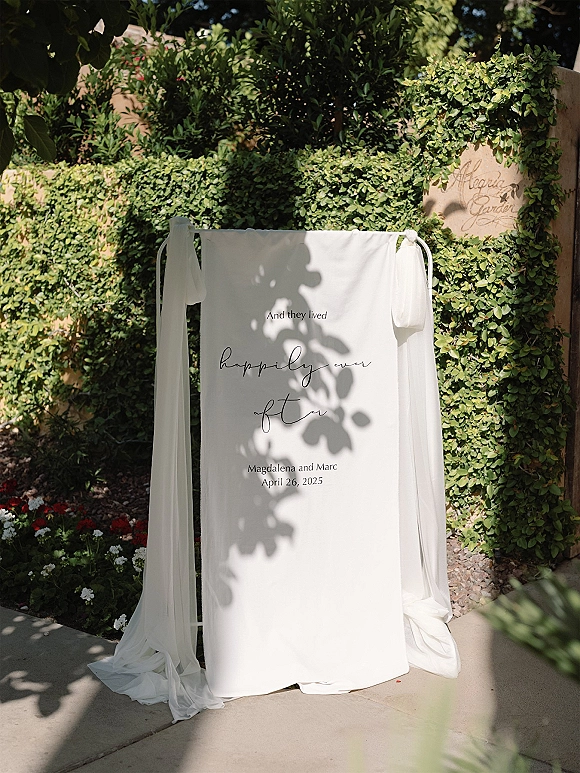 Wedding welcome sign with fabric draped welcome sign styling, calligraphy names and date on a stand against a sunlit hedge wall garden path
