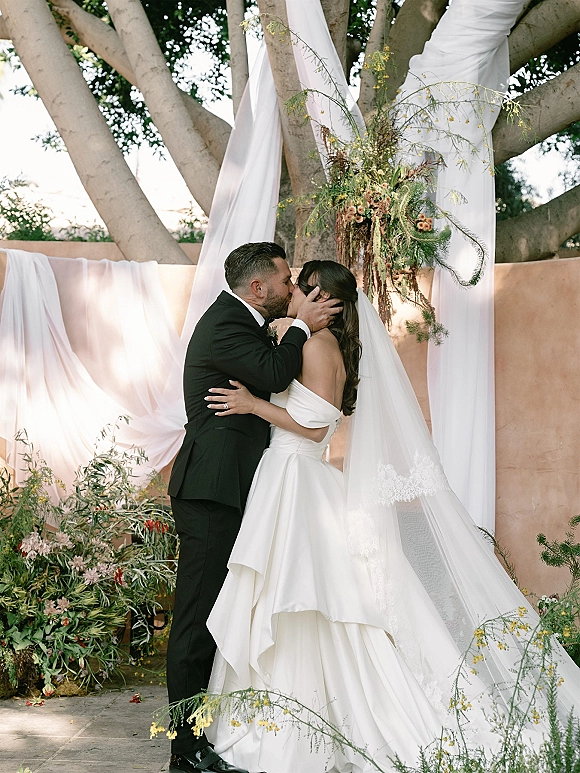 Wedding kiss portrait of bride and groom kissing as her veil blows, framed by floral drapery and greenery in an outdoor courtyard setting