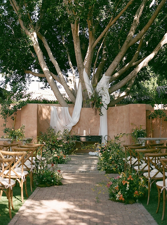 Ceremony setup with wood crossback chairs lining a brick aisle, meadow florals and greenery garlands leading to a white draped altar under a large tree