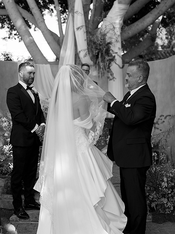 Ceremony moment as father lifts the bride’s lace-edged veil while groom in tux waits before draped greenery under a large garden tree