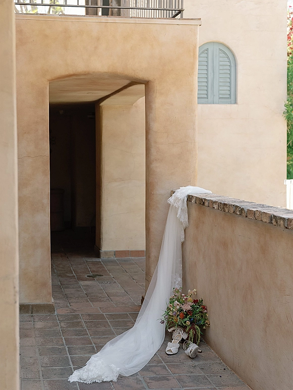 Wedding dress detail with lace wedding dress train draped over a stucco wall, paired with bridal shoes and bouquet in an arched corridor