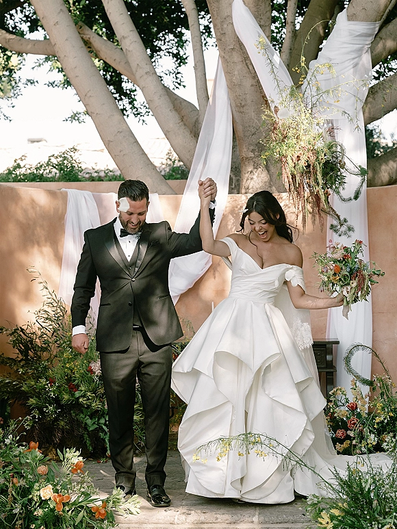 Recessional moment as bride and groom walk down aisle holding hands, bride cheering with bouquet near draped ceremony arch in garden greenery
