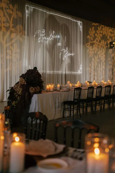 Reception tablescape glowing as a candlelit reception table, with burgundy florals before draped curtains and a wall projection