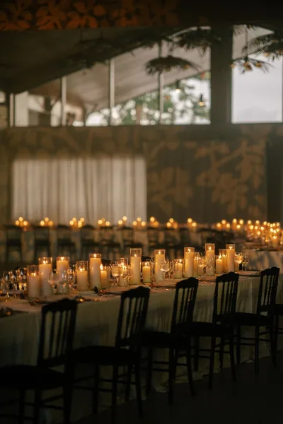 Reception tablescape on a candlelit reception table with clustered glass cylinder vases, set before draped walls and windows.