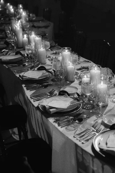 Reception tablescape set as a candlelit wedding table with pillar candles, framed by banquet chairs in a dim room.