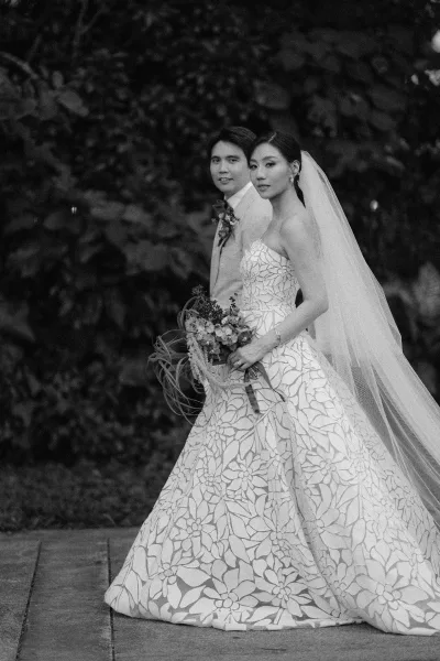 Couple portrait of a bride and groom walking along a garden walkway, her long veil trailing as they pass lush foliage