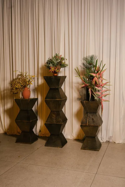 Ceremony flowers create refined altar flower arrangements on dark wood pedestals, with organic shapes set against a simple curtain backdrop.