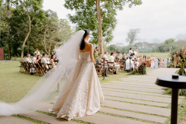 Wedding processional with a bride walking down the aisle, her long veil trailing along a stone path amid garden trees under an open sky.