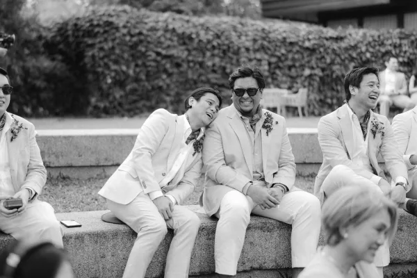 Groomsmen in a black-and-white moment, wearing light suits and sunglasses, laugh together on a stone bench beside hedges and ceremony chairs.