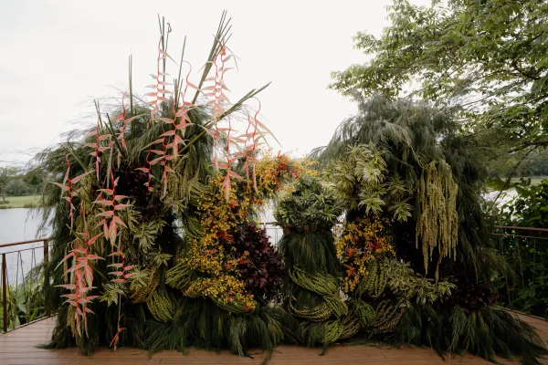 Ceremony backdrop with a tropical wedding altar framed by a greenery ceremony arch, accented by bold heliconia on a lakeside wooden deck.
