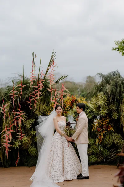 Ceremony moment as the bride and groom hold hands beneath garden trees on a wooden platform, her veil trailing behind her.
