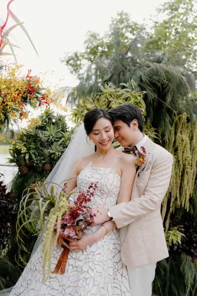 Couple portrait of a bride and groom embrace amid garden greenery, her cascading bridal bouquet trailing as trees and hanging plants frame them.