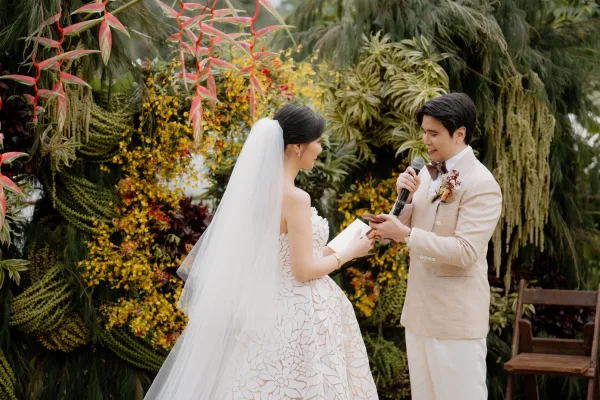 Wedding vows with the groom reading vows to his bride beneath a tropical floral arch amid lush garden greenery.