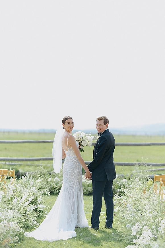 Couple portrait of bride and groom holding hands, bride looking back in veil with white bouquet beside floral aisle chairs in open field