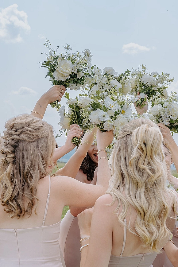 Bridesmaid bouquet moment with bridesmaids holding bouquets, raising white rose and greenery blooms against a bright blue sky backdrop