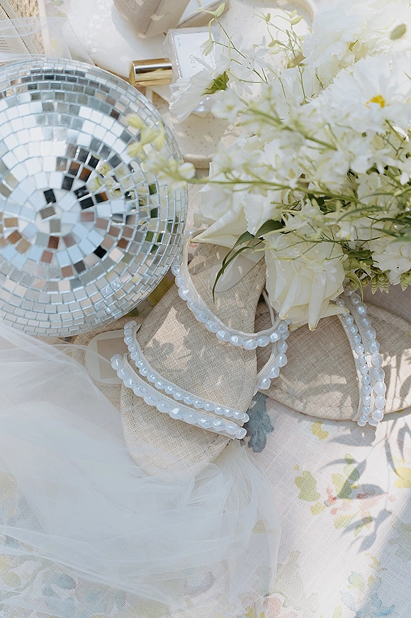 Bridal accessories flatlay of pearl strap bridal shoes with white bouquet, perfume bottle and disco ball on floral linen fabric background