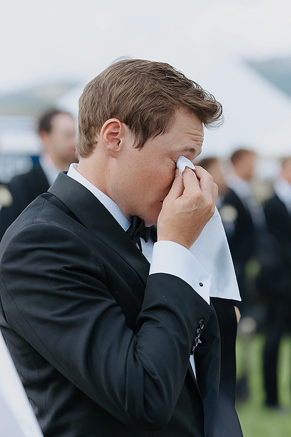 Groom emotional moment, groom crying wedding as he wipes tears with a handkerchief in a black tuxedo, guests blurred on a lawn under open sky