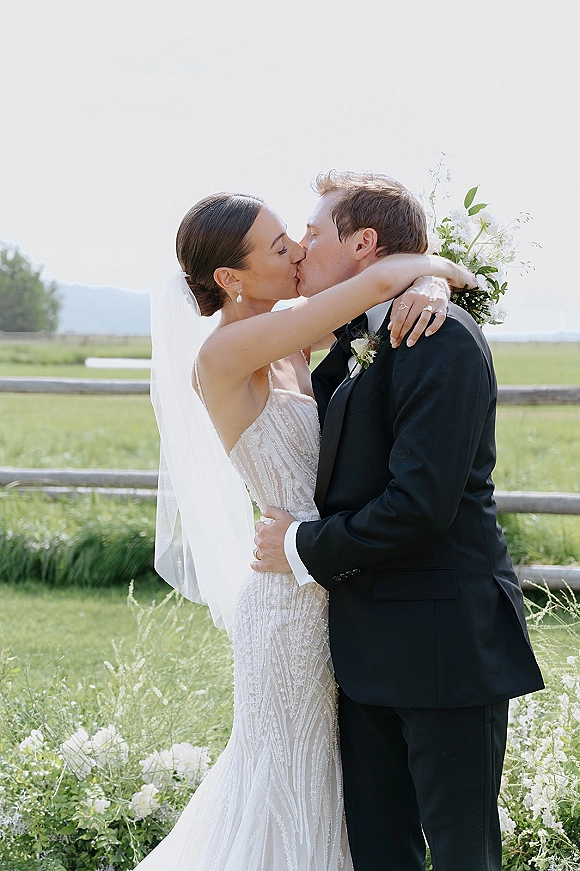 Wedding kiss portrait of bride and groom kissing in a wildflower meadow, her veil and greenery bouquet framed by mountains and a rustic fence